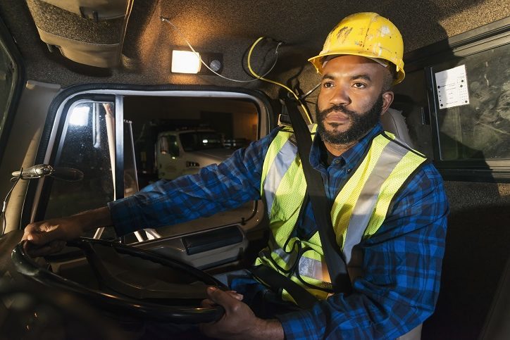 An Afircan American man driving a truck, wearing a hard hat and safety vest.